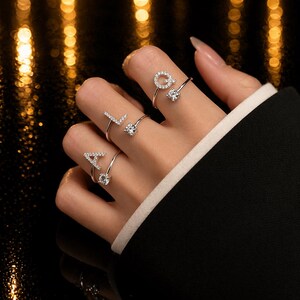Könnte beinhalten: Nahaufnahme einer Hand mit vier silbernen Ringen, die jeweils einen Buchstaben darstellen: A, L und Q. Die Ringe haben ein filigranes Design mit kleinen, funkelnden Edelsteinen. Der Hintergrund ist verschwommen mit goldenem Bokeh.