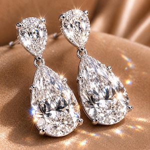 Può includere: Un paio di orecchini pendenti di diamanti scintillanti. Ogni orecchino presenta un diamante a forma di pera, taglio brillante, sospeso da un diamante più piccolo a forma di goccia. Gli orecchini sono incastonati in metallo argentato e riflettono la luce.