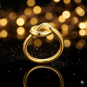 Op de afbeelding: Een goudkleurige ring met een knoopontwerp. De ring is gemaakt van een glanzend, reflecterend metaal. De achtergrond is vervaagd met gouden lichten. Het ontwerp van de ring is een eenvoudige knoop.