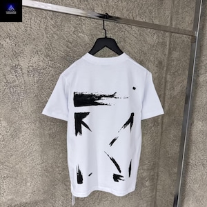 Puede incluir: Camiseta blanca con diseños de pinceladas y flechas negras. La camiseta de manga corta está colgada de una percha negra sobre un fondo neutro. La parte trasera de la camiseta presenta una gran pincelada negra y varias marcas en forma de flecha.