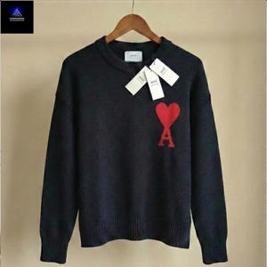 May include: A black crew neck sweater with a red heart and the letter "A" logo. The sweater has long sleeves and a ribbed hem and cuffs. The sweater is hanging on a wooden hanger.