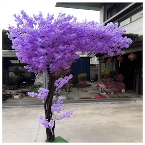 Può includere: Un grande albero artificiale con vibranti fiori viola. L'albero ha un tronco e rami marrone scuro ed è posto su una piccola base verde. Lo sfondo mostra un edificio e altre piante, in un ambiente esterno.