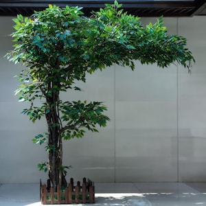 Peut inclure: Un grand arbre artificiel avec des feuilles vertes luxuriantes et un tronc brun. L'arbre est entouré d'une petite clôture en bois. L'arrière-plan est un mur et un sol de couleur claire.