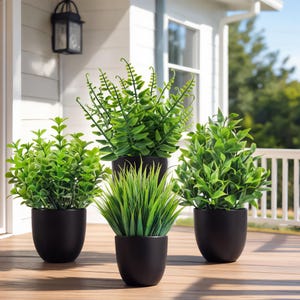 Könnte beinhalten: Vier künstliche Topfpflanzen mit leuchtend grünen Blättern. Die Pflanzen stehen in schwarzen, abgerundeten Töpfen auf einer Holzterrasse. Die Pflanzen weisen verschiedene Texturen und Formen auf und schaffen eine natürliche Ästhetik für die Inneneinrichtung.