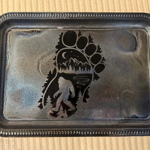 May include: A rectangular metal tray with a Bigfoot design. The tray has a dark, textured surface with a black Bigfoot footprint, silhouette, and mountain scene. The tray's edges are detailed with a decorative border.