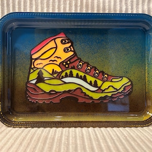 May include: Rectangular rolling tray with a colorful design of a hiking boot. The boot illustration features a mountain landscape with a sunset, trees, and a yellow, green, and brown color scheme. The tray has a blue and yellow gradient background.