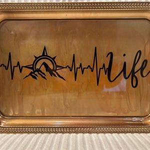 May include: A rectangular gold-colored tray with a decorative border. The tray features a black graphic of a mountain range, compass, and heartbeat line, with the word "life" in cursive script.