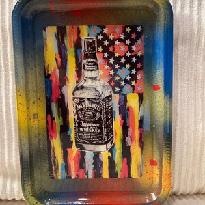 Può includere: Vassoio rettangolare con un design astratto e colorato. Il vassoio presenta una grafica di una bottiglia di whisky Jack Daniel's su uno sfondo che ricorda la bandiera americana. I bordi del vassoio sono dipinti in tonalità di rosso, giallo e blu.