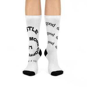 May include: White socks with black heels and toes. One sock has the text "Hustle More Coffee" in a circular design. The other sock has the word "good" repeated in a vertical pattern. The socks are designed for comfort and style.