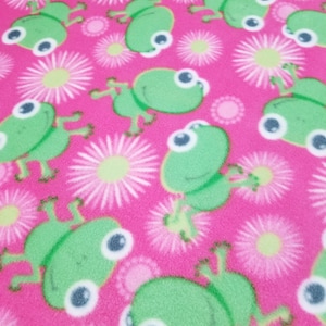 May include: Pink fleece fabric with a repeating pattern of green frogs and white flowers. The frogs have large eyes and are in a jumping pose.