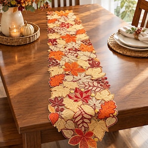 May include: A rectangular table runner featuring an intricate cutwork design of embroidered autumn leaves in shades of orange, red, yellow, and beige, accented with small berry clusters and acorns throughout the detailed foliage pattern.