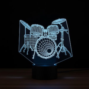 Puede incluir: Una lámpara de noche con forma de batería con efecto 3D azul. La lámpara presenta un diseño de alambre detallado de una batería, que incluye bombo, caja, toms y platillos. La base es oscura y rectangular.