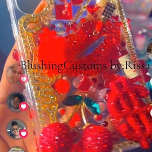 May include: A clear phone case decorated with red rhinestones, a large red lip charm, a pair of red cherry charms, and gold-toned accents. The case features the text Thangya and BlushingCustoms by:Rissa, with various small red and clear embellishments.
