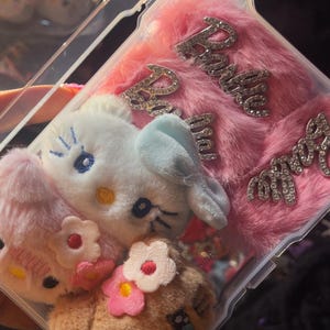 Può includere: Una custodia di plastica trasparente con interno rosa e soffice, ornata da scritte "Barbie" con strass. La custodia è decorata con teste di peluche, tra cui un gatto bianco con un fiocco blu e altri due peluche con accenti floreali.