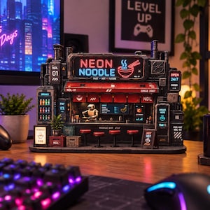 May include: A detailed miniature model of a cyberpunk-style noodle bar featuring a prominent sign that reads NEON NOODLE. The structure includes a small robot figure, red stools, illuminated signage, and various industrial details in black and red tones.
