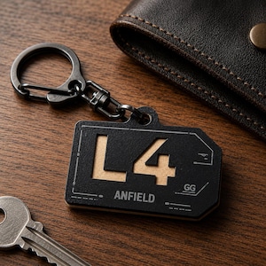 May include: A black rectangular keychain with a metallic finish featuring the cutout text L4 and the printed word ANFIELD. The keychain includes a dark metal split ring and a swivel clasp, showcasing technical line details on its surface.