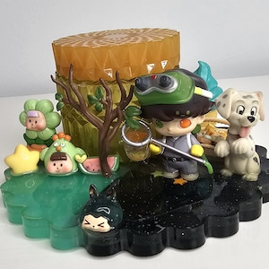 May include: A colorful figurine set featuring a central honey-colored structure with a textured top. The base is a dark, scalloped shape. Various cartoon figures, including a dog, are arranged around the base.