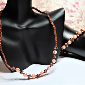 May include: A copper-colored necklace and bracelet set. The necklace features a woven chain with alternating peach-colored and dark brown beads. The bracelet mirrors the design with peach and brown beads. The jewelry is displayed on a black mannequin.