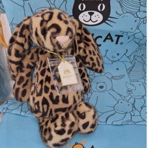 Puede incluir: Un conejito de peluche con estampado de leopardo. El conejito tiene largas orejas caídas, una nariz blanca y una pequeña etiqueta adjunta. El fondo es una tela azul claro con ilustraciones de gatos y osos de dibujos animados.