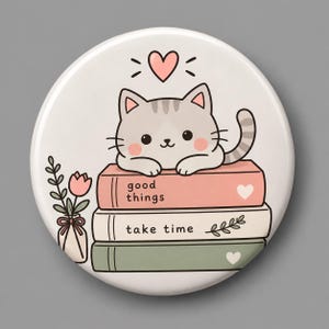 May include: A round pinback button featuring a cartoon gray tabby cat resting on a stack of three books. The top book is pink with the text good things, the middle is white with take time, and the bottom is green. A pink heart and flower are included.