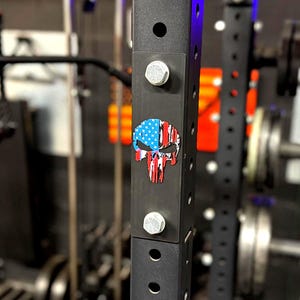 Puede incluir: Un rack de pesas negro con una pegatina de calavera con la bandera estadounidense. La pegatina es roja, blanca y azul. El rack tiene pernos y agujeros para ajustes. Otros equipos de gimnasio son visibles en el fondo.