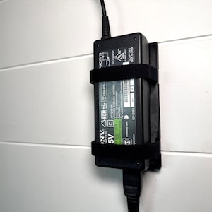 May include: A black Sony adapter, secured to a white wall with a black strap. The adapter has a green and white label with text including "5V" and "ADP-75UB A". A black power cord extends from the bottom.