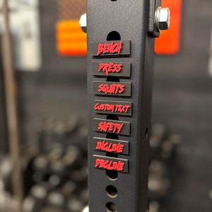 Peut inclure: Gros plan sur un rack de musculation noir avec des étiquettes rouges. Les étiquettes indiquent "Bench Press", "Squats", "Custom Text", "Safety", "Incline" et "Decline". Le rack est percé de trous et fixé avec des boulons.