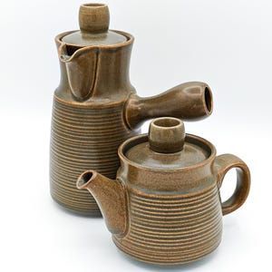 May include: A set of two brown ceramic vessels with a ribbed texture. The taller vessel features a side handle and a spout, while the shorter vessel includes a lid with a knob, a small spout, and a side handle. Both items have a dark brown glaze.