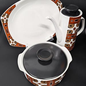 May include: A ceramic dinnerware set featuring a white serving platter, a tall coffee pot, and a lidded casserole dish. Each piece is decorated with a repeating geometric pattern in burnt orange, black, and white, with solid black lids.