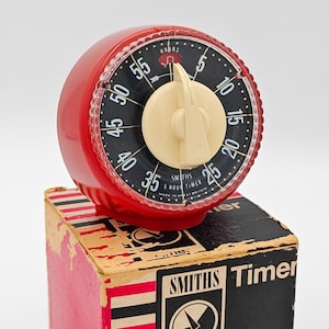 May include: A round red Smiths 5 hour timer with a cream-colored dial knob and a black face featuring white numbers from 5 to 55. The timer sits atop its original cardboard box, which is printed with the brand name Smiths and the word Timer.