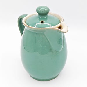 May include: A sage green ceramic pitcher with a matching lid featuring a small knob handle and a central steam vent. The pitcher has a curved handle on the side, a wide pouring spout, and a smooth, glossy finish with light cream-colored trim.