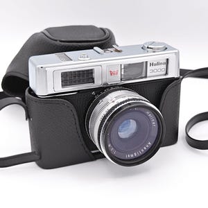 May include: A silver and black Halina 3000 film camera with a textured black leather case and a matching shoulder strap. The camera features a prominent lens labelled Anastigmat 1:2.8 and a top plate with the brand name and a red crown logo.