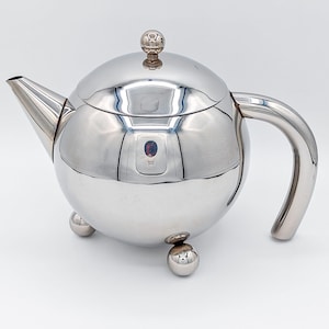 Puede incluir: Una tetera esférica de metal plateado pulido con un pico curvo, un asa a juego y una tapa extraíble con un pequeño pomo esférico. La tetera se apoya sobre tres pequeñas patas esféricas, lo que le confiere un aspecto equilibrado y elevado.
