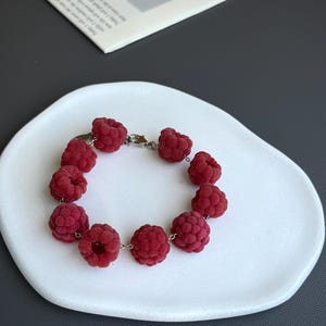 May include: A raspberry-themed bracelet composed of multiple red raspberry-shaped beads strung together with silver-colored chain links. The bracelet is displayed on a white, organically shaped plate against a gray background.