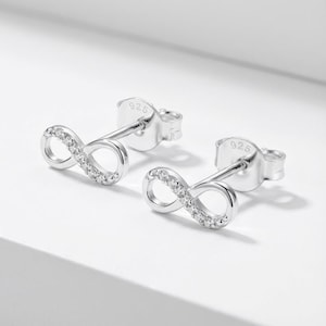 May include: A pair of silver-colored infinity symbol stud earrings. Each earring features a small infinity symbol with a row of clear, sparkling stones. The earrings are set against a white background.