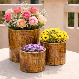 May include: Three ceramic pots with a textured, brown finish, each filled with colorful flowers. The largest pot holds pink and peach roses with white filler flowers. The other two pots contain yellow daisies and purple flowers.