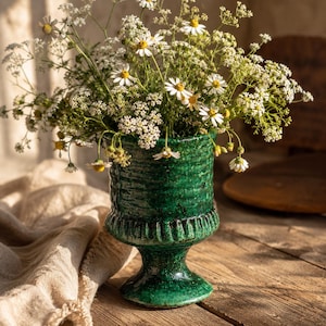 Könnte beinhalten: Eine grüne Keramikvase mit strukturierter Oberfläche, gefüllt mit einem Strauß weißer und gelber Wildblumen. Die Vase hat einen Sockel und einen breiten Rand. Die Blumen blühen in voller Blüte, mit kleinen weißen Blütenblättern und gelben Zentren. Die Vase steht auf einer Holzoberfläche.