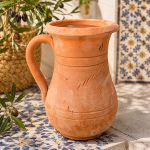 Può includere: Una brocca in terracotta con corpo arrotondato, bordo largo e un unico manico curvo. La superficie presenta linee orizzontali incise e sottili intagli a forma di foglia. L'argilla ha una calda tonalità arancione terrosa con variazioni.