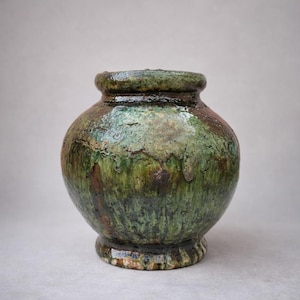 Puede incluir: Un jarrón de cerámica hecho a mano con un cuerpo redondeado y una superficie texturizada y vidriada. El jarrón presenta una paleta de colores verdes y marrones, con un aspecto ligeramente áspero y envejecido. Tiene una pequeña base decorativa y un cuello estrecho.