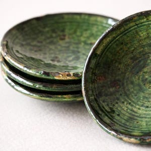 May include: A stack of four small, round, green ceramic plates. The plates have a rustic, handmade appearance with a textured surface and a dark rim. The plates are a deep, earthy green color.