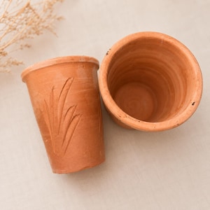 May include: Two terracotta cups with a natural reddish-brown finish. One cup is positioned upright showing a hollow interior, while the other is tilted to display a carved leaf-like pattern on its exterior surface. Both items are unglazed.