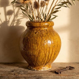 Puede incluir: Un jarrón de cerámica texturizada de color amarillo mostaza, con un cuerpo bulboso y un cuello estrecho. El jarrón está lleno de flores y tallos secos. Una llave antigua descansa sobre la superficie de madera frente al jarrón.