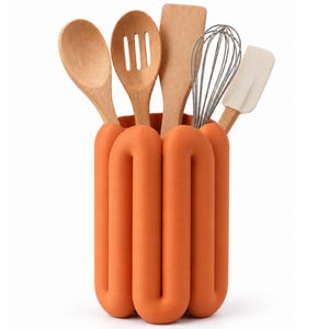 Puede incluir: Un soporte cilíndrico para utensilios de color naranja mate con un diseño tubular continuo, que contiene cinco herramientas de cocina: una cuchara de madera, una cuchara ranurada de madera, una espátula de madera, un batidor de metal y una espátula de silicona color crema.