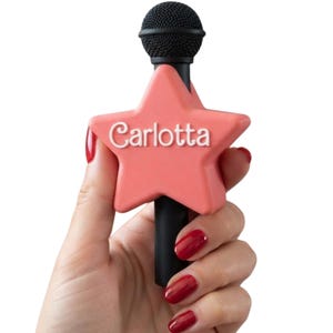 Pode incluir: Um microfone de mão preto com um acessório em forma de estrela na cor coral no cabo. A estrela tem o nome Carlotta em relevo com letras brancas em estilo cursivo na parte frontal. A cabeça do microfone é preta com um acabamento em rede.