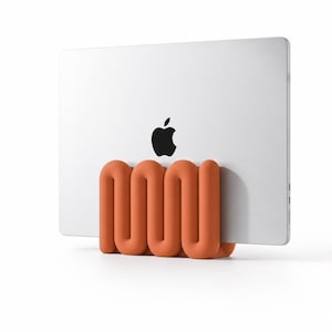 Könnte beinhalten: Ein silberner Laptop mit einem schwarzen Apple-Logo wird von einem matten, terrakottafarbenen Ständer aufrecht gehalten. Der Ständer hat ein durchgehendes, abgerundetes, schlangenförmiges oder zickzackförmiges Design, das das Gerät stützt.