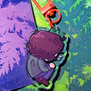 Op de afbeelding: Een doorzichtige acryl sleutelhanger met een cartoonillustratie van een persoon met donkerpaars haar, gekleed in een grijs jasje en een blauwe broek, zittend in een opgerolde houding. De sleutelhanger is bevestigd aan een oranje plastic karabijnhaak.