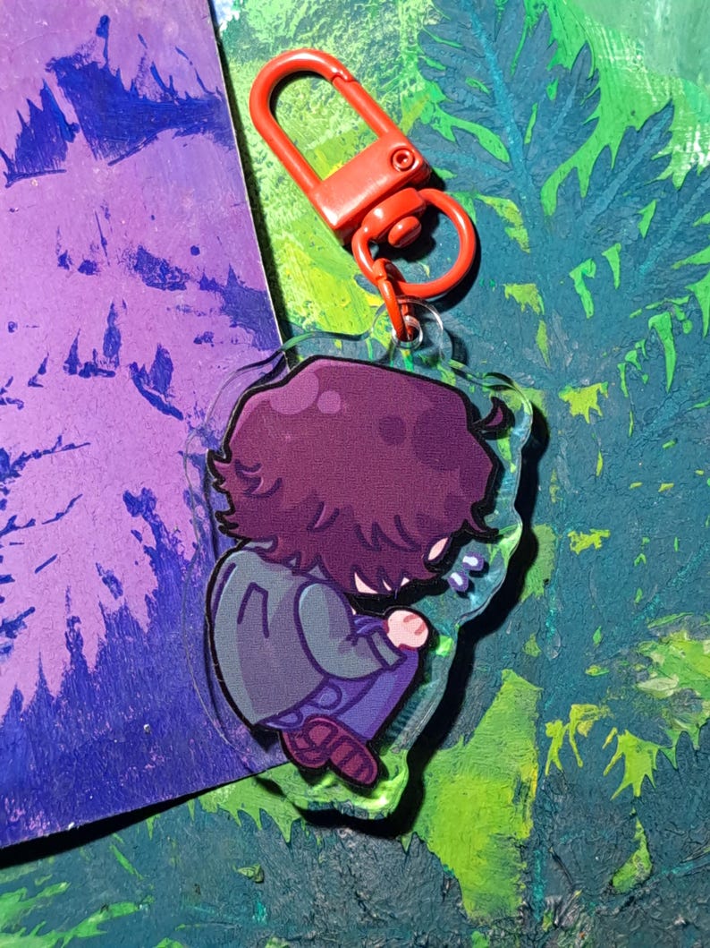 Op de afbeelding: Een doorzichtige acryl sleutelhanger met een cartoonillustratie van een persoon met donkerpaars haar, gekleed in een grijs jasje en een blauwe broek, zittend in een opgerolde houding. De sleutelhanger is bevestigd aan een oranje plastic karabijnhaak.