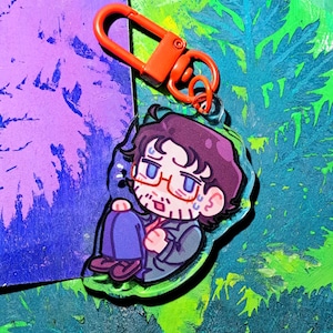 Op de afbeelding: Een doorzichtige acryl sleutelhanger met een cartoonillustratie van een persoon met donker haar, een bril en een baard, gekleed in een donker jasje en een blauwe broek, zittend in een opgerolde houding, voorzien van een feloranje plastic clip.
