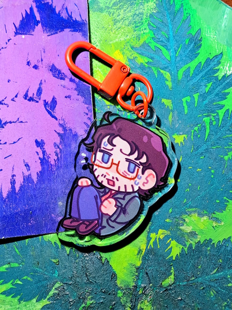 Op de afbeelding: Een doorzichtige acryl sleutelhanger met een cartoonillustratie van een persoon met donker haar, een bril en een baard, gekleed in een donker jasje en een blauwe broek, zittend in een opgerolde houding, voorzien van een feloranje plastic clip.