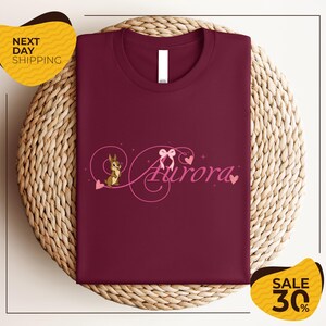 a burgundy t-shirt with a pink and purple design, including a cartoon character and the word "Aurora". The t-shirt is displayed on a woven basket, and there are two yellow sale signs in the background.